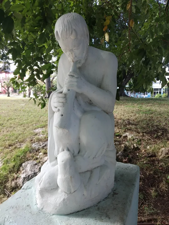Escultura de piedra blanca en un parque: un pastor sentado toca una flauta con un cabrito a sus pies, bajo las ramas de un árbol