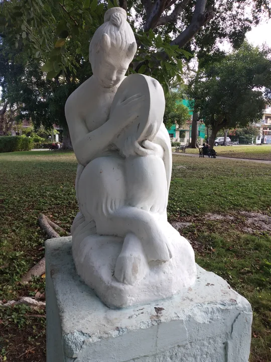 Escultura de piedra blanca en el mismo parque: una mujer sentada, con el pelo recogido, abraza contra el pecho un pandero