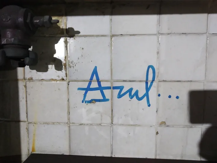 La palabra «Azul…» escrita a mano con rotulador sobre los azulejos blancos de una pared, junto a una vieja llave de paso