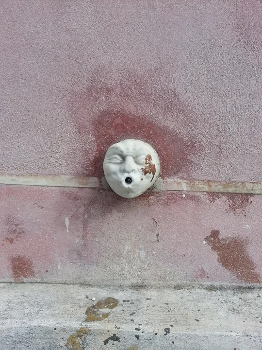 Un petit visage de faïence blanche, yeux fermés et bouche ouverte, encastré dans un mur rose écaillé