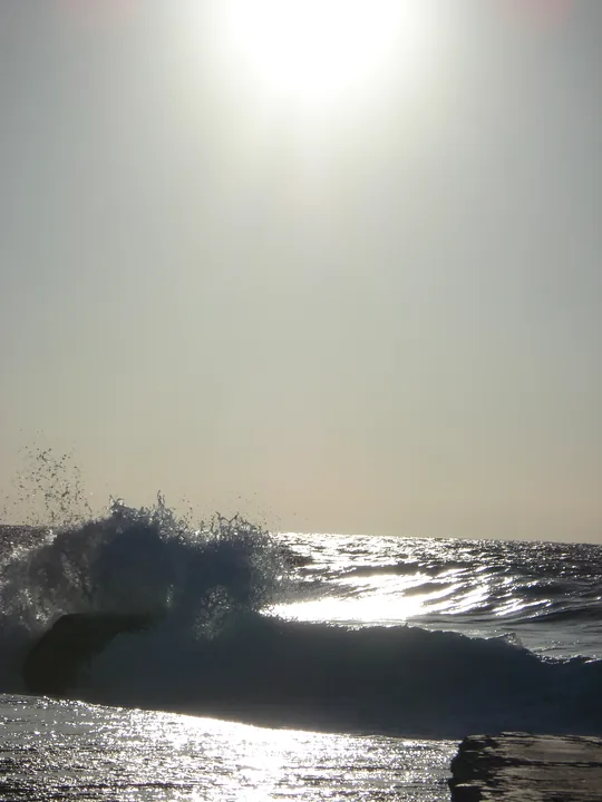 Le soleil de face au-dessus de la mer, une vague éclatant à contre-jour sur les rochers