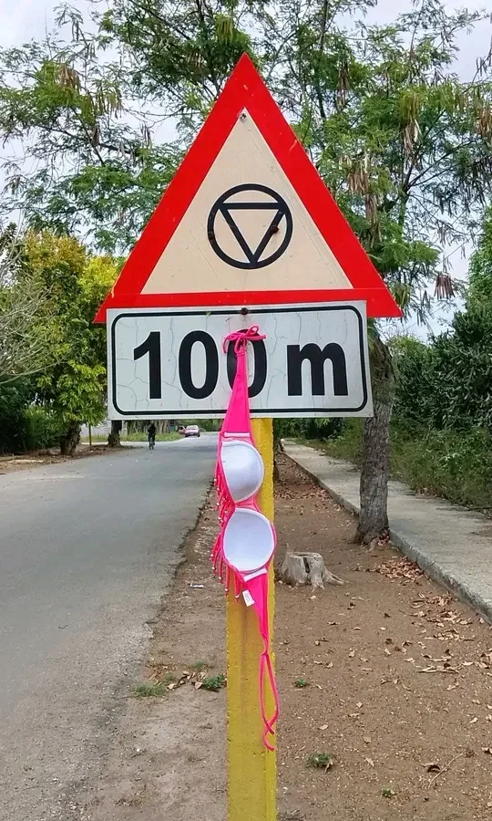 A triangular road sign with a 100 m plate, a pink bikini top hanging from the post
