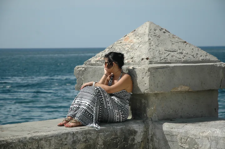 Une femme assise sur le mur du Malecón, à côté d'un bloc de béton, la mer derrière elle